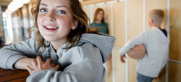 En högstadietjej står i en skolkorridor och tittar in i kameran med ett leende