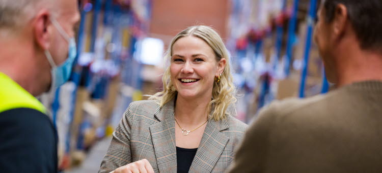 En person ler mot kameran, i förgrunden två personer med ryggen mot kameran, i bakgrunden lagerhyllor. 